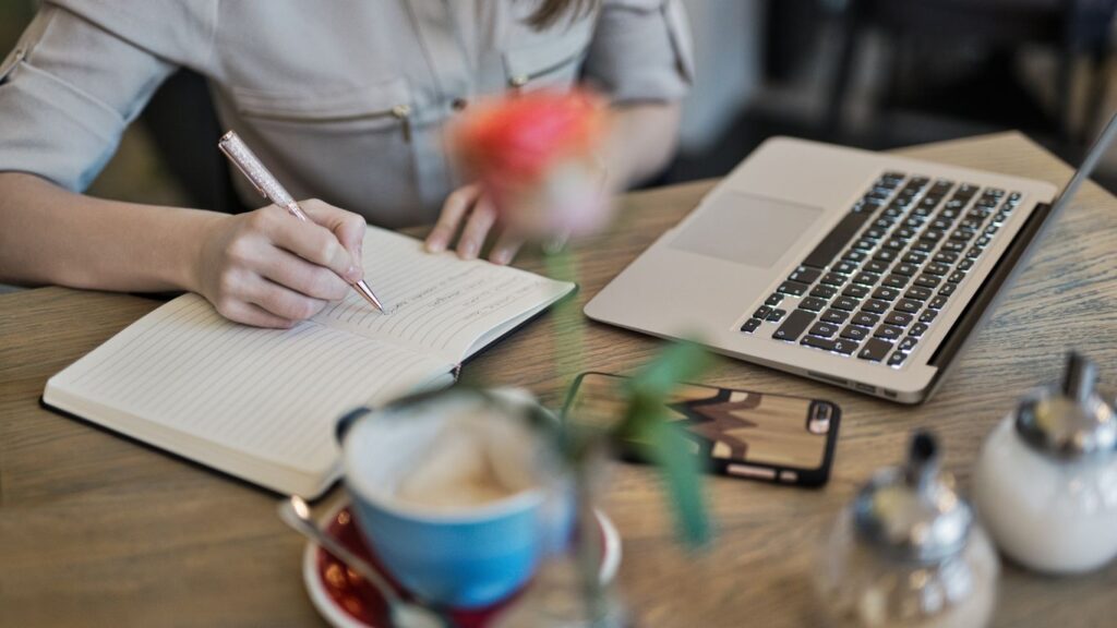 A person writing in a notebook with a rose gold pen while sitting at a table with a laptop, smartphone, and coffee cup.